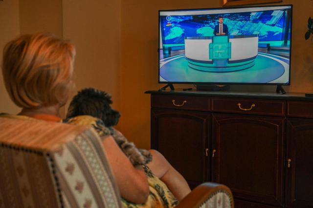 A woman sits with a dog on an armchair to watch on the TV the new news service of the relaunched state-run TV channel 1 on August 20, 2026, in Budapest, Hungary, after the news service resumed following a 44-day forced break. (Photo by Attila KISBENEDEK / AFP)