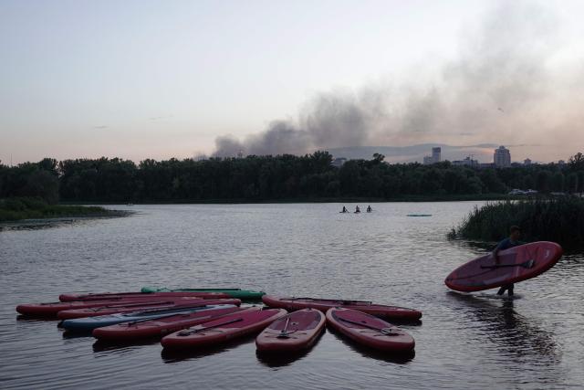 A man carries a canoe along the Dnipro River as smoke from a fire following a Russian air strike rises in the background in Kyiv on August 20, 2026, amid the Russian invasion of Ukraine. (Photo by Genya SAVILOV / AFP)