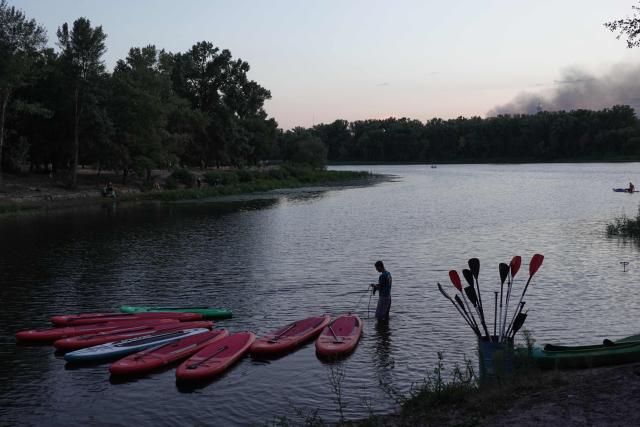A man holds a rope linked to canoes along the Dnipro River as smoke from a fire following a Russian air strike rises in the background in Kyiv on August 20, 2026, amid the Russian invasion of Ukraine. (Photo by Genya SAVILOV / AFP)