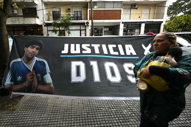(FILES) A woman walks past a banner depicting Argentine football legend Diego Maradona and demanding justice outside the court before a trial hearing on his death in San Isidro, Buenos Aires province, Argentina on June 9, 2026. The trial over the death of football legend Diego Maradona has been postponed for a week, until August 27, to verify whether incorrect medical tests were used in the expert reports, the court announced on August 20, 2026. (Photo by Luis ROBAYO / AFP)