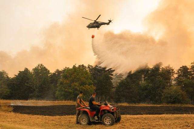 People watch as a Serbian Armed Forces helicopter drops water on a wildfire in Deliblatska Pescara (Deliblato Sands), near the village of Grebenac, some 70 kilometres northeast of Belgrade on August 20, 2026. Firefighters were battling wildfires across the Balkans as days of high temperatures and drought conditions continue to drive blazes in several countries, including Serbia. Scientists have long warned that climate change caused by human use of fossil fuels will make periods of drought more intense and longer-lasting, creating the ideal conditions for wildfires. (Photo by Uros Arsic / AFP)