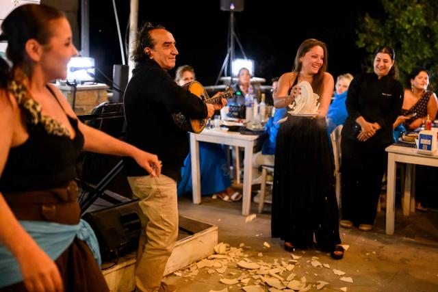 Konstantin Kostanoglu (C) and Tuba Demir (L) perform at a tavern during the Feast of the Dormition of the Virgin Mary, on Gokceada (Imroz) Island, Çanakkale Province of Turkey, on August 14, 2026. Hundreds of Greeks return from Athens and around the world to Imbros each August 15 to celebrate the Virgin Mary. Like Istanbul and nearby Tenedos, the island was exempt from the 1923 population exchange between Greece and Turkey, which displaced more than 1.5 million people. (Photo by Yasin AKGUL / AFP)