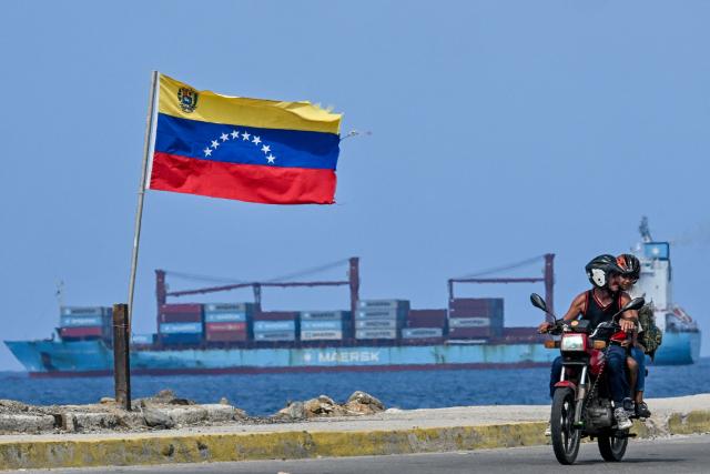 A man rides a motorbike next to a waving Venezuelan flag as a cargo ship sails at La Guaira Port, Venezuela on August 22, 2026. The Venezuelan Parliament announced an investigation into opposition lawmaker Antonio Ecarri on August 22, 2026, and removed him from his legislative post after he proposed dollarizing the economy with the advice of US economist Steve Hanke. (Photo by Juan BARRETO / AFP)