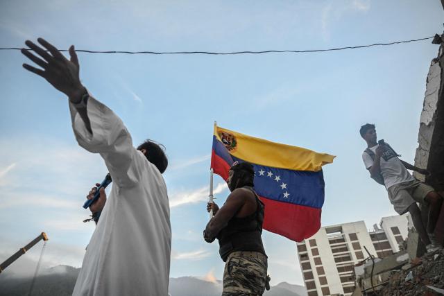 A person waves a Venezuelan flag as a priest holds a mass with residents and relatives of the earthquake victims from the state-run OPP housing blocks two months after the 7.2 and 7.5-magnitude earthquakes in La Guaira, Venezuela on August 24, 2026. The magnitude 7.2 and 7.5 tremors on June 24, 2026, devastated the coastal state of La Guaira, where at least 190 buildings fell apart. Venezuela's interim government reported 6,509 deaths on August 24, 2026, exactly two months after the twin earthquakes. (Photo by Maryorin Mendez / AFP)