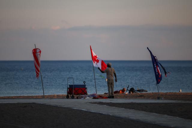 A man stands next to a Canadian flag at dusk on a beach at Lake Ontario in Toronto, Ontario, on August 27, 2026. US President Donald Trump announced that he is renaming Lake Ontario -- a large body of water shared by the United States and Canada -- "Lake America." Trump signed an order in the White House in a move that comes amid a spiraling trade war between the world's largest economy and its northern neighbor. (Photo by Cole Burston / AFP)
