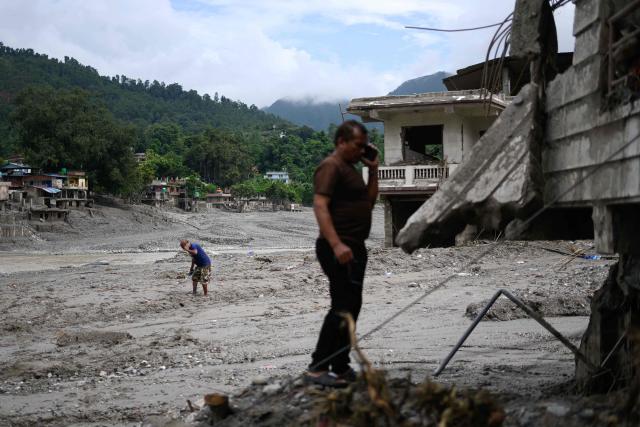 Men talk on their phones amidst  houses heavily covered in mud in the aftermath of flash floods at Devighat in Nepal's Nuwakot district on August 29, 2026. Nearly 3,000 people were still missing on August 29 and 633 confirmed dead after flash floods and landslides hit the Nepal-Tibet border. (Photo by Manan VATSYAYANA / AFP)
