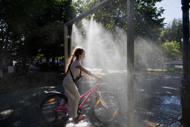 A girl rides a bicyle under a water sprinkler in Berehove on August 28, 2026, amid the Russian invasion of Ukraine. (Photo by Tetiana DZHAFAROVA / AFP)