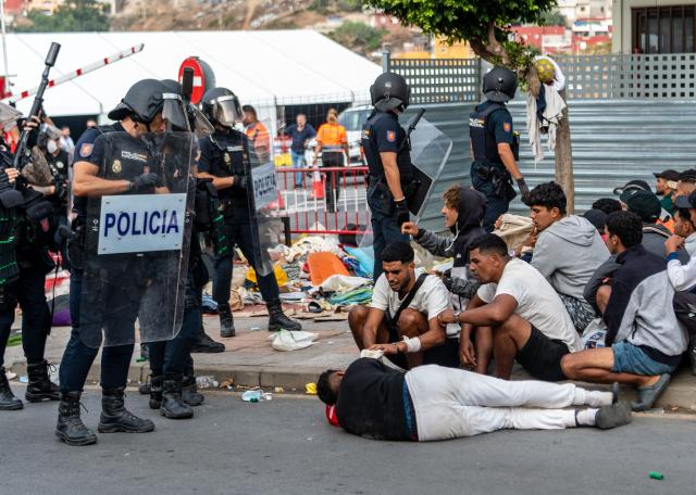 A migrants lays on the ground as Spain's National Police officers stand in front of migrants waiting on the sidewalk before being registered in Loma Colmenar district of Ceuta on August 31, 2026. Tensions remain high in the territory of 80,000 inhabitants since tens of thousands of people crossed into Ceuta from Morocco over several days in July, triggering a Europe-wide migrant alert. Spanish Prime Minister said the rush of migrants to the Spanish enclave was still under investigation, though he ruled out any responsibility on the part of Moroccan authorities. (Photo by Antonio Sempere / AFP)