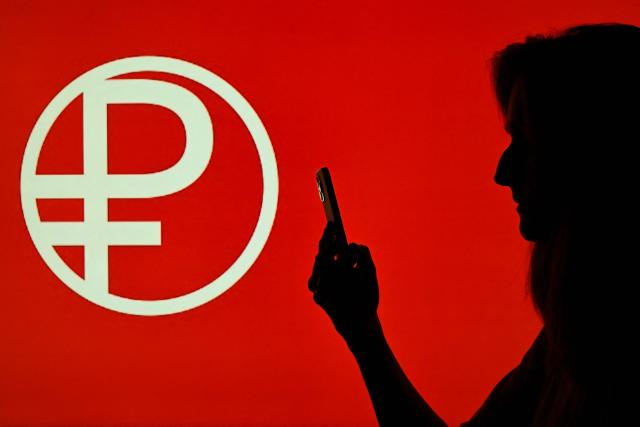 This illustration photograph shows a person holding a smartphone, while standing in front of a screen displaying the Digital Ruble logotype, pictured in Moscow on August 31, 2026. A law will take effect on September 1, 2026 spurring the widespread adoption of the digital ruble. Digital rubles will be stored in special accounts for individuals and organisations, which can be opened on the Bank of Russia platform. Access to digital ruble accounts will be possible through familiar remote channels: bank mobile apps and online banking. (Photo by Igor IVANKO / AFP)