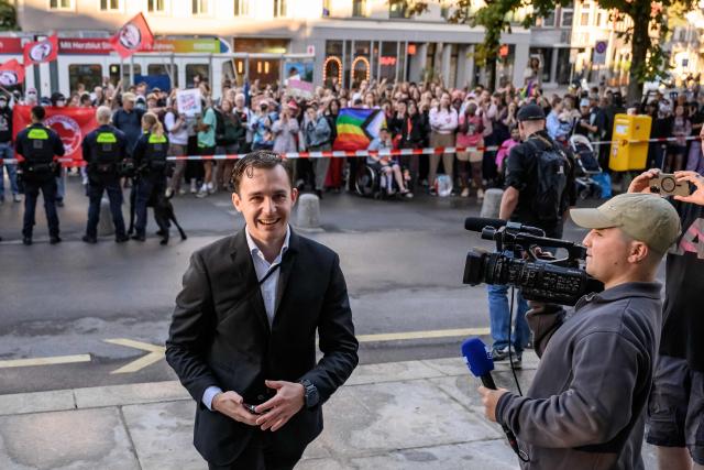 Manuel Corchia, a leading figure of the Swiss far-right group Junge Tat, smiles as he arrives at Zurich District Court for his trial, with LGBT and antifa protesters standing behind him, in Zurich, Switzerland, on September 4, 2026. Six people from the far-right group Junge Tat appeared before the Zurich District Court, accused of hatred-related offenses, coercion, rioting, property damage and trespassing. The group's two main leaders face up to six months in prison. (Photo by Fabrice COFFRINI / AFP)