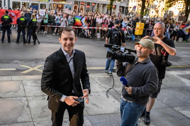 Manuel Corchia, a leading figure of the Swiss far-right group Junge Tat, smiles as he arrives at Zurich District Court for his trial, with LGBT and antifa protesters standing behind him, in Zurich, Switzerland, on September 4, 2026. Six people from the far-right group Junge Tat appeared before the Zurich District Court, accused of hatred-related offenses, coercion, rioting, property damage and trespassing. The group's two main leaders face up to six months in prison. (Photo by Fabrice COFFRINI / AFP)