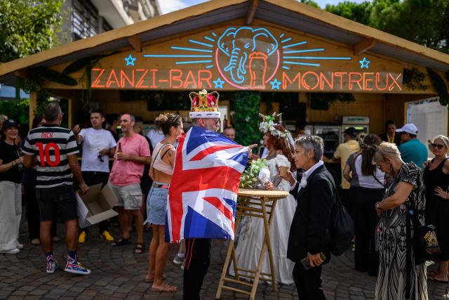 A fan wearing a crown and a Union Jack flag gestures at a bar during Montreux Freddie Day, marking the 80th anniversary of the birth of the Queen frontman Freddie Mercury, in Montreux, Switzerland, on September 5, 2026. (Photo by Fabrice COFFRINI / AFP)