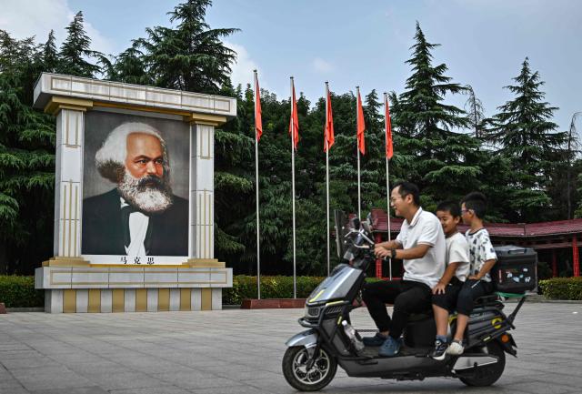 This picture taken on August 20, 2026, shows people riding past a portrait of the philosopher and theorist of socialism Karl Marx in the main "East is Red Square" of Nanjie Commune, in central China's Henan province. In the ruling Communist Party's telling, Mao remains the revolutionary hero -- his face on banknotes, his portrait overlooking Tiananmen Square, and his body preserved in a mausoleum. (Photo by Pedro PARDO / AFP) / TO GO WITH: China-Mao-anniversary, by Sam DAVIES