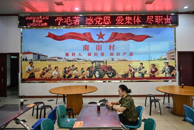 This picture taken on August 20, 2026, shows a woman checking her phone in front of a socialist inspired mural at a restaurant in Nanjie Commune, in central China's Henan province. In the ruling Communist Party's telling, Mao remains the revolutionary hero -- his face on banknotes, his portrait overlooking Tiananmen Square, and his body preserved in a mausoleum. (Photo by Pedro PARDO / AFP) / TO GO WITH: China-Mao-anniversary, by Sam DAVIES