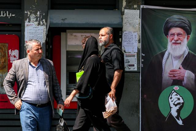 A banner depicting Iran's late supreme leader Ali Khamenei is displayed along a Bank Mellat automated teller machine (ATM) as people walk past, in Tehran on September 7, 2026. Iran's top negotiator threatened on September 6 of a more forceful response to any further US attacks after Tehran targeted American warships in their latest round of clashes. The exchanges of fire, which began with US raids last week, come amid a deadlock in the six-month war between the two foes. (Photo by AFP) / 