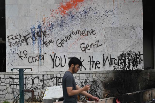 (FILES) A man uses a phone as he walks past a graffitied wall on the facade of the torched Parliament building in Kathmandu on September 14, 2025, days after the anti-corruption Gen-Z protests that left structure in flames. Reeling from devastating floods that cut a swath of destruction through the Himalayan nation, Nepal marks the first anniversary of the September 2025 mass youth-led anti-corruption protests that toppled the previous government. (Photo by Pedro PARDO / AFP)