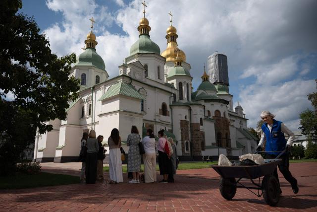 Visitors listen to a tour guide while an employee pushes a cart in front of the St. Sophia Cathedral in Kyiv on September 11, 2026, amid the Russian invasion of Ukraine. Despite the constant threat of missile and drone attacks, restorers have uncovered three compositions hidden from view for more than 70 years at Saint Sophia Cathedral, an architectural monument of Kyivan Rus. The former cathedral is one of the city's best known landmarks and the first heritage site in Ukraine to be inscribed on the World Heritage List. (Photo by Genya SAVILOV / AFP)