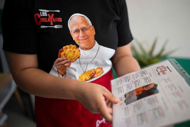 A worker displays a menu featuring dishes named in honor of Pope Leo XIV at a restaurant specializing in traditional Chiclayo cuisine in Lima on September 15, 2026, while wearing a T-shirt depicting Pope Leo XIV holding "Tortitas de Choclo.". Pope Leo XIV will visit Uruguay, Argentina and Peru from November 6 to 17, the Vatican said on August 5, in a return to the continent where the pontiff spent decades as a missionary. (Photo by Ernesto BENAVIDES / AFP)