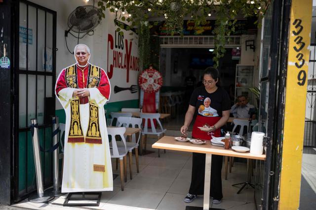 A worker serves dishes at a restaurant specializing in traditional Chiclayo cuisine in Lima on September 15, 2026, while wearing a T-shirt depicting Pope Leo XIV holding "Tortitas de Choclo.". Pope Leo XIV will visit Uruguay, Argentina and Peru from November 6 to 17, the Vatican said on August 5, in a return to the continent where the pontiff spent decades as a missionary. (Photo by Ernesto BENAVIDES / AFP)