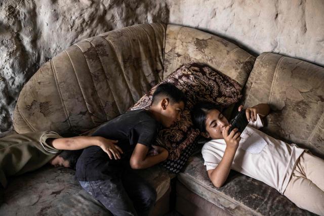 TOPSHOT - Palestinian boy Waheed (C) watches his sister play video games inside a cave that their family has turned into their home in the village of Al-Tuwani, in the Israeli-occupied West Bank, on September 11, 2026. After his home was destroyed by Israeli forces in 2023, Mohammed Fadel Amr took shelter in caves, where he now lives with his two wives, Fatma Amr and Arwa Amr, and their eleven children. Mohammed and his family say they face almost daily harassment from settlers living in a nearby illegal Israeli settlement outpost. At the end of August 2026, Israeli forces reportedly ransacked the family's makeshift home during a night raid, and settlers later stole his goats, depriving him of his livelihood. (Photo by John WESSELS / AFP)
