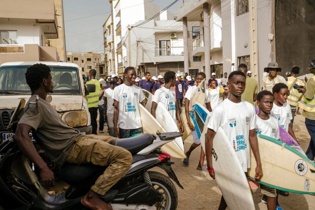 Young people walk with their surf board as they follow a torch bearer carrying the Youth Olympic Flame during the flame's 46-day tour in Dakar on September 17, 2026. The Youth Olympic Flame will travel across Senegal's 14 regions before returning to Dakar for the opening ceremony on October 31, 2026. Senegal will host the Summer Youth Olympic Games from October 31 to November 13, 2026, bringing together 2,700 athletes competing in 25 sports. (Photo by Carmen Abd Ali / AFP)