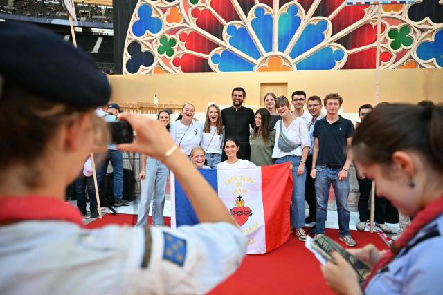 Young Catholics pose with a priest, holding a French tricolour flag bearing the Sacred Heart of Jesus and the inscription "Coeur Sacré de Jésus", while scouts in the foreground photograph them ahead of the Pope's arrival at the Stade de France stadium, in Saint-Denis on the outskirts of Paris on September 25, 2026 to lead a prayer vigil with young people, as part of his four days visit to France. (Photo by LOU BENOIST / AFP)