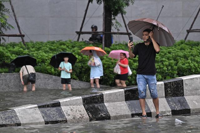 A man takes pictures as people walk through flood waters along Phetchaburi Road in Bangkok on September 26, 2026. Bangkok authorities declared on September 26 a disaster across all 50 districts of the Thai capital after continuous rain caused heavy flooding. (Photo by Lillian SUWANRUMPHA / AFP)