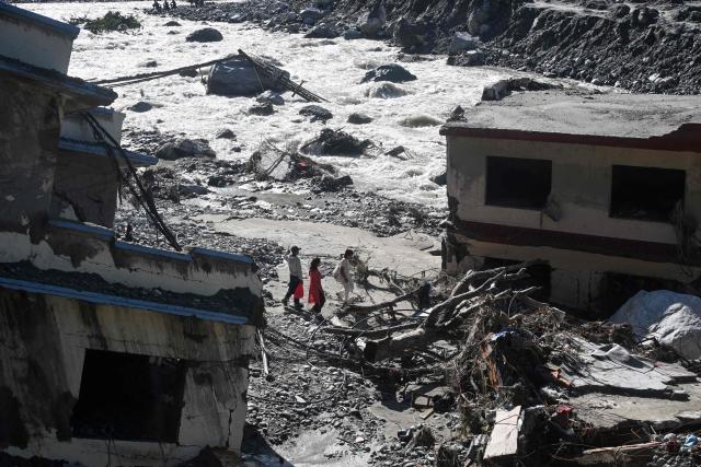 People walk past damaged houses covered in sludge in the aftermath of the August 26 flash flood, at Betrawati Bazaar in Nepal's Rasuwa district on September 28, 2026. The Himalayan nation suffered one of the worst catastrophes in its history late last month when a tsunami-like flood, triggered by a high-altitude glacial mountain collapse along the Nepal-China border, cut a swathe of destruction claiming more than 1,400 lives and leaving 5,000 unaccounted for. (Photo by Prakash MATHEMA / AFP)