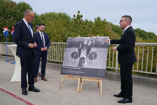 German Chancellor Friedrich Merz (L) and the mayor of Saarlouis Marc Speicher (R) stand next to an iconic historic photograph (depicting then French President François Mitterrand (L) and then German Chancellor Helmut Kohl holding hands during a reconciliation ceremony on September 22, 1984, in Douaumont, near Verdun, in northeastern France) as they inaugurate streets in honor of the late former German Chancellor Helmut Kohl and the late former French President Francois Mitterrand, in Saarlouis, western Germany on September 28, 2026.  (Photo by Andreas Schlichter / AFP)