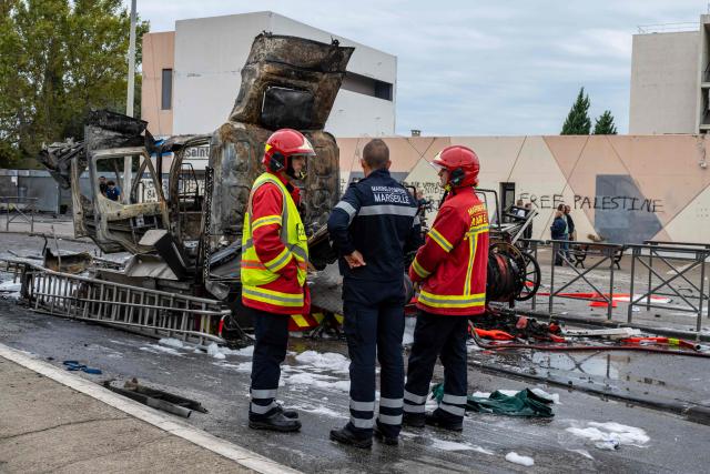 Firefighters stand in front of a burnt firefighter truck at the entrance of the Saint-Exupéry high school amid high school student protests, in the 15th arrondissement of northern Marseille, southern France, on October 1, 2026. Since late September, students have staged protests outside schools over grievances ranging from timetables, class sizes and a shortage of teachers to overheated classrooms during this year's heatwaves. The protests started in the Ile-de-France region around Paris but have now spread to other cities nationwide. (Photo by Miguel MEDINA / AFP)