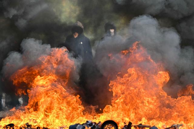TOPSHOT - Protesting students stand in front of a burnt barrier during   a blockading action outside the secondary school Le Corbusier, in Aubervilliers, northern Paris on October 1, 2026. Since late September, students have staged protests outside schools over grievances ranging from timetables, class sizes and a shortage of teachers to overheated classrooms during this year's heatwaves. The protests started in the Ile-de-France region around Paris but have now spread to other cities nationwide. (Photo by LOU BENOIST / AFP)