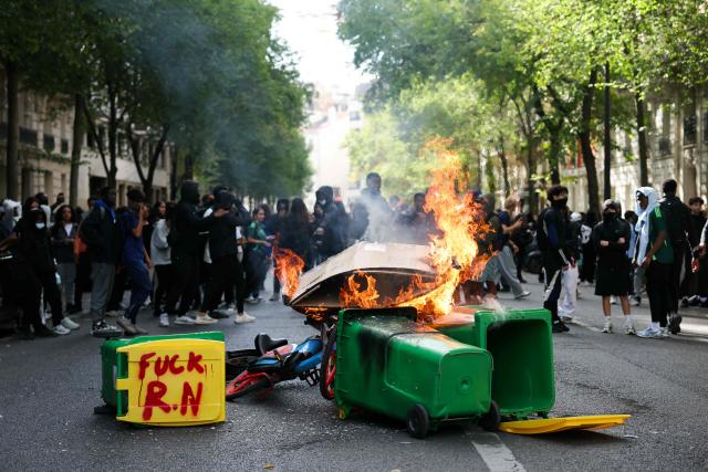 Students look at bins burning with a sentence reading "Fuck RN (Rassemblement National)" as protesting students carry out blockading action near place de la Nation, in Paris, on October 2, 2026. Since late September, students have staged protests outside schools over grievances ranging from timetables, class sizes and a shortage of teachers to overheated classrooms during this year's heatwaves. The protests started in the Ile-de-France region around Paris but have now spread to other cities nationwide. (Photo by Thomas SAMSON / AFP)