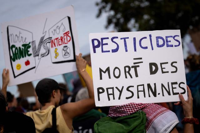Protesters hold placards reading "Our health vs your interests" and "Pesticides = death of farmers" during a demonstration as part of an international day of mobilisation against the Syngenta industrial site, which produces pesticides, to denounce the agrochemical industry and demand its shutdown, in Saint-Pierre-la-Garenne, Normandy, on October 3, 2026. (Photo by Guillaume SALIGOT / AFP)