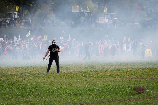 A protester wearing a face covering stands amid smoke as other demonstrators and tractors gather behind during a demonstration as part of an international day of mobilisation against the Syngenta industrial site, which produces pesticides, to denounce the agrochemical industry and demand its shutdown, in Saint-Pierre-la-Garenne, on October 3, 2026. (Photo by Guillaume SALIGOT / AFP)
