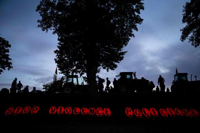 Protesters and tractors are silhouetted behind a banner reading "Stop police violence" during a demonstration as part of an international day of mobilisation against the Syngenta industrial site, which produces pesticides, to denounce the agrochemical industry and demand its shutdown, in Saint-Pierre-la-Garenne, Normandy, on October 3, 2026. The demonstration is called by Terre de Liens Normandie association, an initiative of the farmers' union Confederation paysanne, the Cancer Colere awareness group, the CSVPO (support collective for victims of pesticides in the West) and the Soulevements de la terre and gather farmers, farm workers, sick people and residents of contaminated territories. (Photo by Guillaume SALIGOT / AFP)