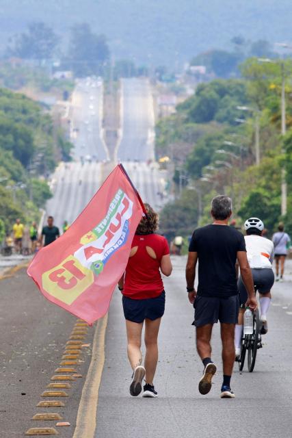 A supporter of Brazil's President and re-election candidate for the leftist Workers' Party (PT) Luiz Inacio Lula da Silva carries a campaign flag while walking along a road during the presidential election in Brasilia on October 4, 2026. Brazilians head to the polls in a highly polarized presidential election between incumbent leftist President Luiz Inacio Lula da Silva -seeking a fourth presidential mandate- and right-wing senator Flavio Bolsonaro, who emerged as the leading figure of the conservative movement following the conviction and imprisonment of his father, former president Jair Bolsonaro. (Photo by Igo Estrela / AFP)