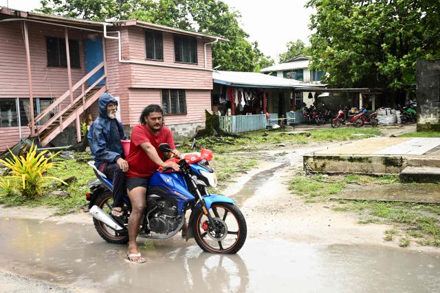 This photo taken on October 6, 2026 shows a Tuvaluan man and pillion rider waiting with their motorcyle on a waterlogged street in Funafuti, Tuvalu. (Photo by Saeed KHAN / POOL / AFP)