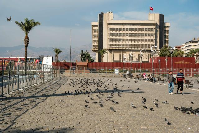 Pedestrians walk past pigeons and police fences as Turkish Police block Konak square due to potential protests after Izmir mayor joined the ruling Justice and Development Party (AKP) in Izmir, on October 7, 2026. The mayor of Izmir, Turkey's third city and an opposition stronghold, on Wednesday defected to the ruling AKP, in a huge symbolic victory for Turkish President Recep Tayyip Erdogan. Mayor Cemil Tugay's decision to join the Islamic-rooted Justice and Development Party (AKP) was announced by Erdogan at a parliamentary group meeting in Ankara where he personally welcomed the mayor in a party-like atmosphere broadcast live on TV. (Photo by Murat Kocabas / AFP)