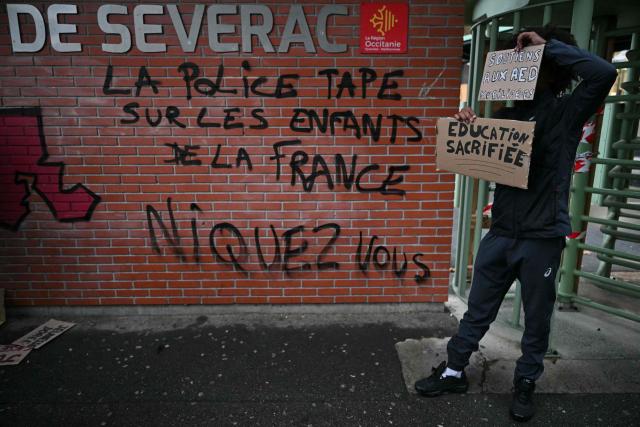 A high-school student protester poses with two placards reading "Support for teaching assistants" (up) and "Education Sacrificed" in front of the entrance wall with a graffiti reading "The police are cracking down on France's children" during a blockade of high-school Deodat de Severac in Toulouse, on October 8, 2026, as part of a nationwide protest movement over learning conditions. Since late September, pupils have barred the entrances to hundreds of schools, complaining of teacher absences, overloaded timetables, dilapidated classrooms and buildings ill-adapted to rising temperatures, among other grievances. (Photo by Lionel BONAVENTURE / AFP)