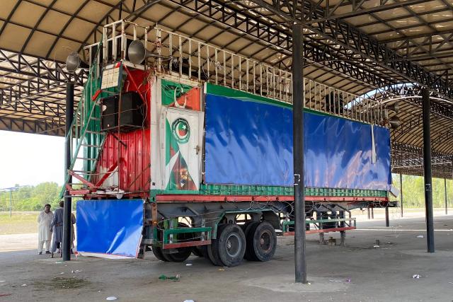 People stand next to an abandoned container truck which was used by inactive Khyber Pakhtunkhwa chief minister Muhammad Sohail Afridi during a protest march organised by the Pakistan Tehreek-e-Insaf (PTI) party to demand the release of jailed former prime minister Imran Khan, at a bus terminal in Mardan on October 12, 2026. Pakistan placed a northern province under emergency rule on October 11, the president's office said, following anti-government demonstrations by supporters of jailed former prime minister Imran Khan and militant violence in the northwest. (Photo by AFP)