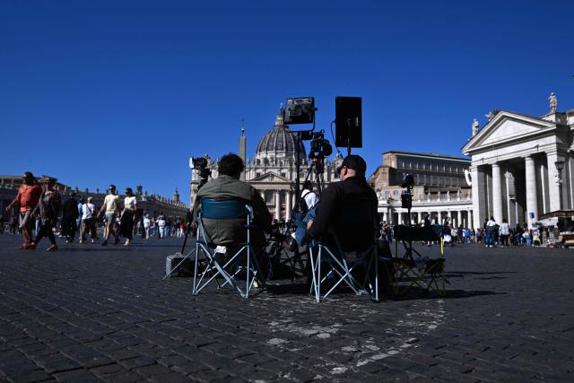 Men sit behind a camera and lights for broadcast TV at St Peter's square in the Vatican on October 12, 2026. Pope Leo XIV has said on October 11, 2026 he will have an operation to remove a lump on his lung this week, the first time a pope has made a personal medical announcement of this kind. (Photo by Tiziana FABI / AFP)