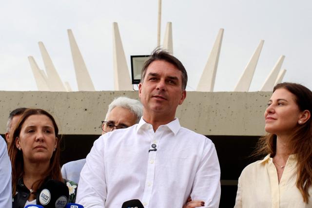 Brazil's right-wing Senator and presidential candidate for the Liberal Party (PL) Flavio Bolsonaro (C) speaks to the media alongside his wife Fernanda Bolsonaro (R) following a mass marking the celebration of Brazil's patron saint, Our Lady of Aparecida, at the Metropolitan Cathedral of Brasilia in Brasilia, Brazil, on October 12, 2026. (Photo by Sergio Lima / AFP)