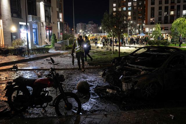 Local residents watch a Ukrainian firefighters working by wreckage cars in front of a high-rise residential building damaged following a Russian drone attack, in Kyiv on October 12, 2026, amid the Russian invasion of Ukraine. (Photo by Genya SAVILOV / AFP)