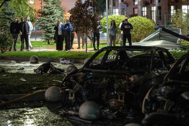 Local residents stand behind burned vehicles in front of a high-rise residential building damaged following a Russian drone attack, in Kyiv on October 12, 2026, amid the Russian invasion of Ukraine. (Photo by Genya SAVILOV / AFP)