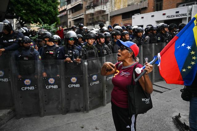 A woman holding a national flag shouts slogans in front of members of the police during a march by active and retired workers from health and education unions, and relatives of political prisoners demanding salary increase and freedom for political prisoners, near the Miraflores presidential palace, in Caracas. on October 13, 2026. (Photo by Juan BARRETO / AFP)