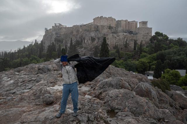 A tourist tries to put on a rain jacket the Areopagus Hill, as the Acropolis is seen in the background, during a rain shower in Athens, on October 14, 2026. (Photo by Angelos TZORTZINIS / AFP)