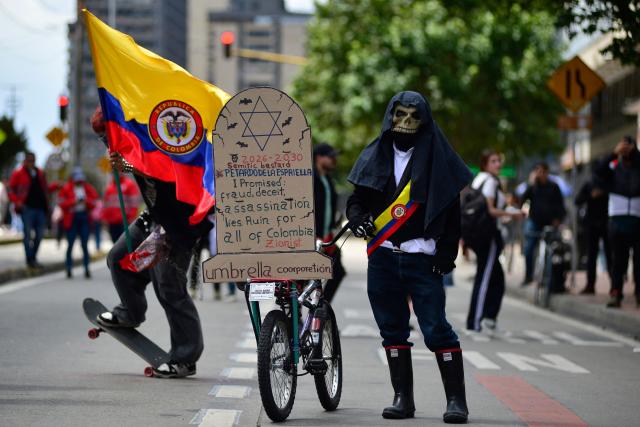 A demonstrator wearing a skull mask and draped in a Colombian presidential sash stands beside a bicycle carrying a placard with anti-government messages during a protest against the government of Colombian President Abelardo de la Espriella in Bogota on October 14, 2026, as part of a nationwide day of demonstrations called by labor unions, teachers' organizations, student groups and opposition sectors. (Photo by Sebastian BARROS / AFP)