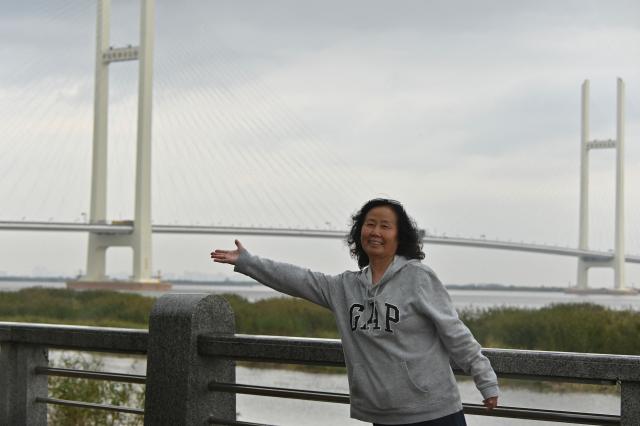 A woman poses for a photo with the new cross-border bridge linking China and North Korea in the background in Dandong, Liaoning Province, on October 15, 2026. A new four-lane road bridge linking China and North Korea finally opened on October 15, 12 years late, enabling the Asian giant and its isolated nuclear-armed neighbour to step up cross-border trade substantially. (Photo by Michael ZHANG / AFP)