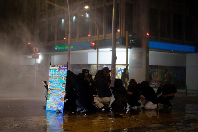 Demonstrators take cover from a police water cannon during clashes following a protest against Colombian President Abelardo de la Espriella in Bogotá, Colombia, on October 14, 2026. Thousands of Colombians took to the streets of cities across the country on October 14, 2026, to protest the policies of new right-wing President Abelardo de la Espriella. (Photo by Pierre BRIOULT / AFP)
