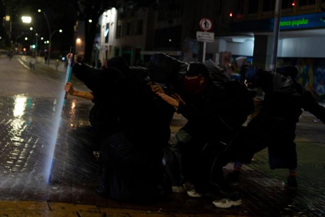 Demonstrators take cover from a police water cannon during clashes following a protest against Colombian President Abelardo de la Espriella in Bogotá, Colombia, on October 14, 2026. Thousands of Colombians took to the streets of cities across the country on October 14, 2026, to protest the policies of new right-wing President Abelardo de la Espriella. (Photo by Pierre BRIOULT / AFP)